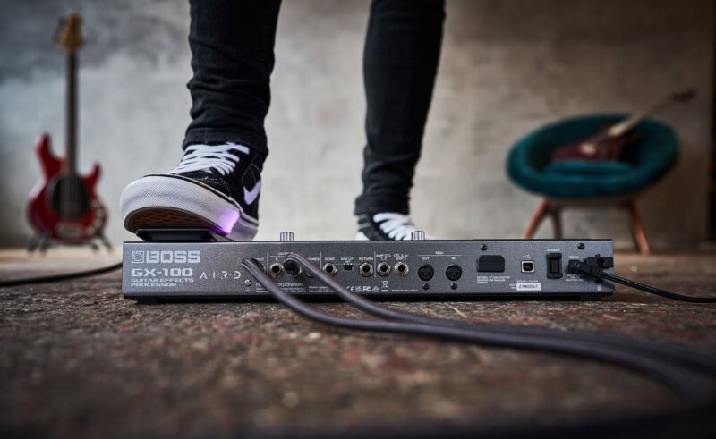 Guitar effects processor and footswitch in a music studio with a person wearing black sneakers, close-up of the back panel showing input/output ports and power connection.