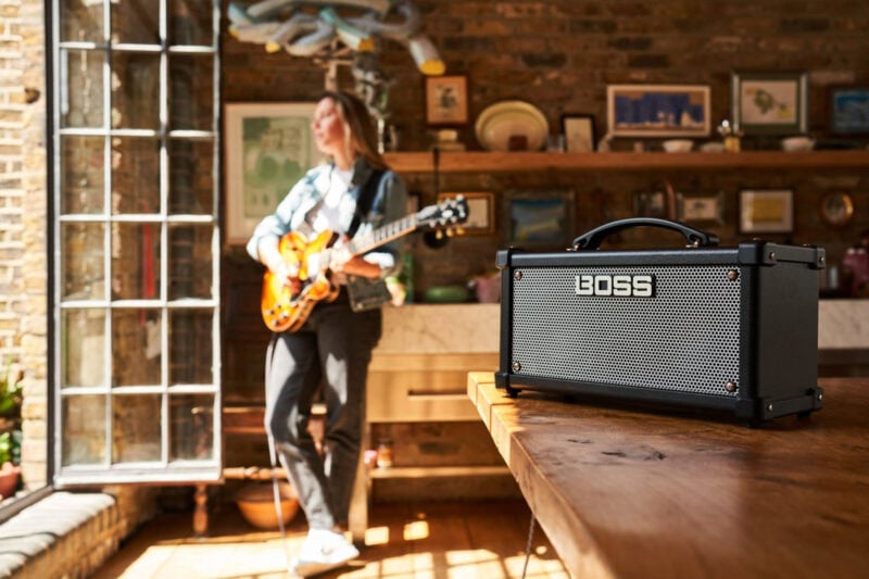 Bright BOSS guitar amplifier on wooden table in a cozy music shop, with a woman playing an electric guitar in the background.