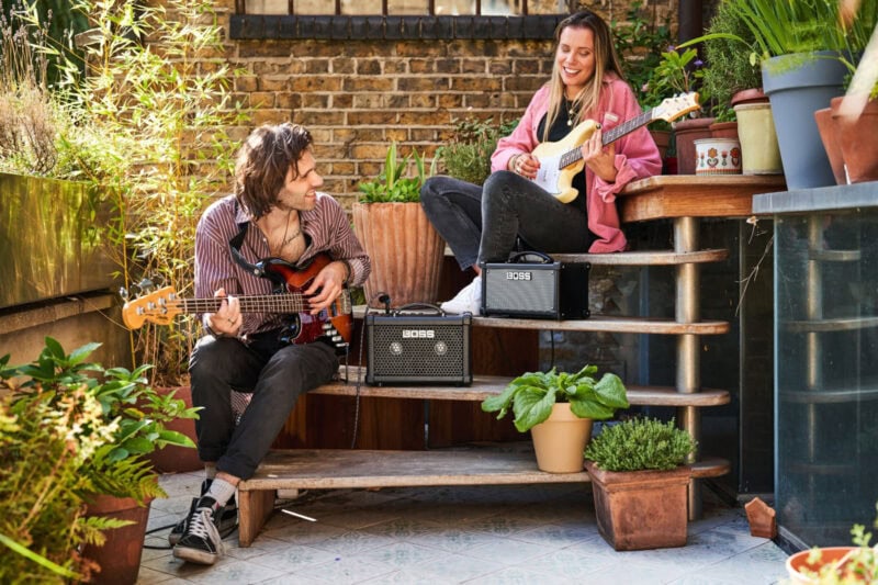 Lively outdoor scene with two young adults playing electric guitars and enjoying music on a sunny terrace surrounded by potted plants and greenery.