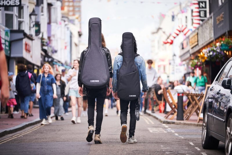 Guitar cases carried by two women walking through a busy street in Swords Dublin filled with shops, people, and outdoor dining, capturing a lively urban environment.