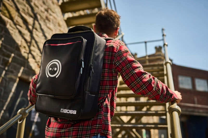 Back view of a young man in a red tartan shirt with a black BOSS backpack, standing outdoors near a staircase, with a clear blue sky and brick building in the background.