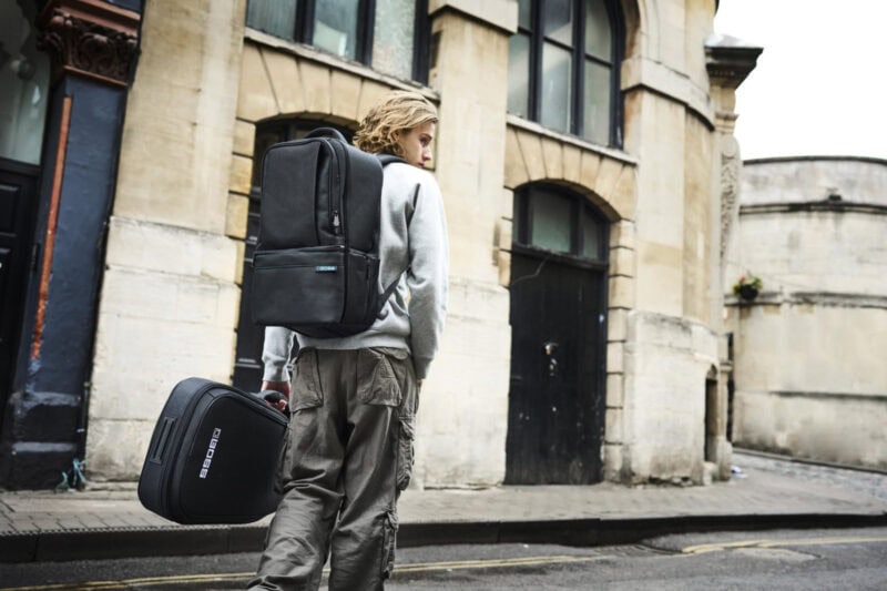Dark grey backpack and gig bag carried by a young man walking past a historic building on a city street.