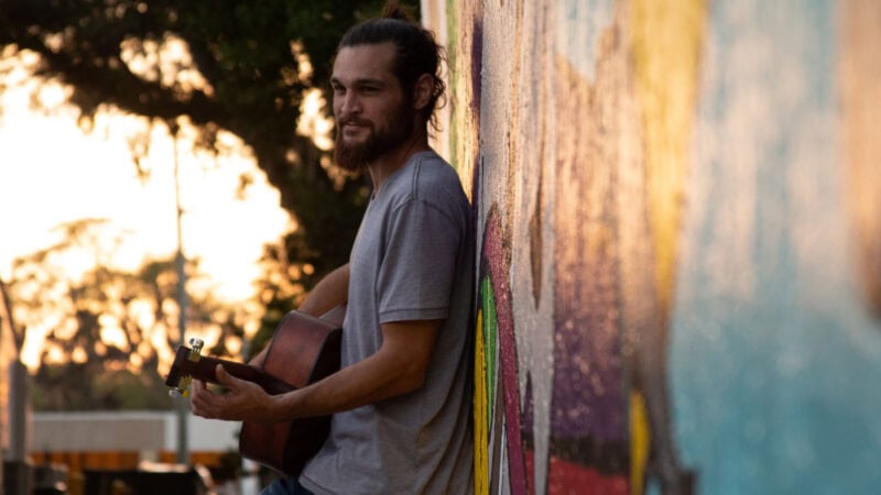 A young man playing an acoustic guitar outdoors at sunset, leaning against a colourful mural wall, creating a relaxed and artistic atmosphere in Swords, Dublin.