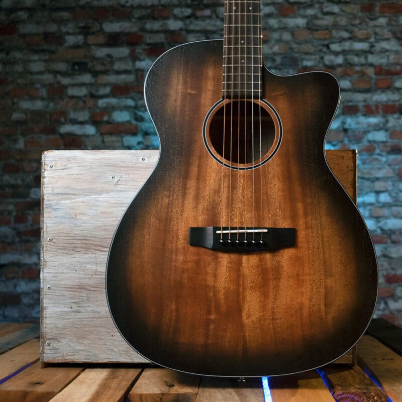 Rich mahogany acoustic guitar with black pickguard on wooden table against exposed brick wall.