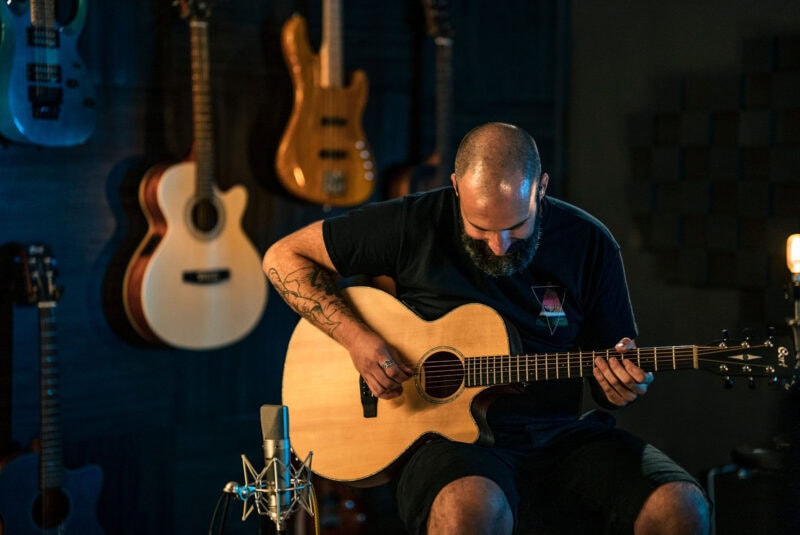 High-angle shot of a bearded man with tattoos playing an acoustic guitar in a music studio with several guitars hanging on the dark blue wall behind him.