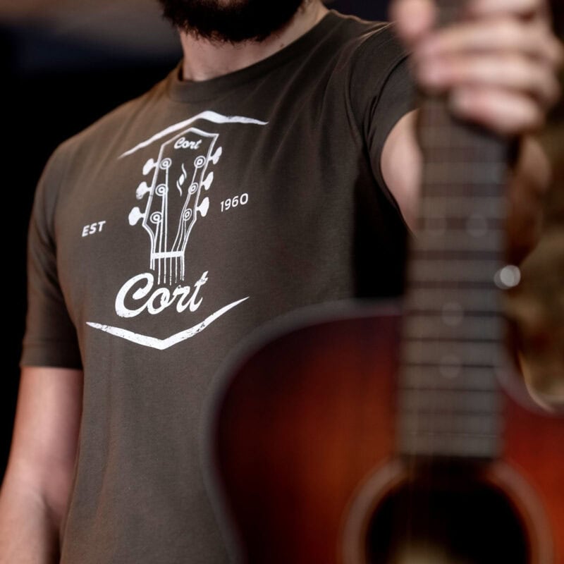 Vintage-style graphic t-shirt with guitar logo and "Cort" text, worn by a person holding an acoustic guitar in a music shop setting.