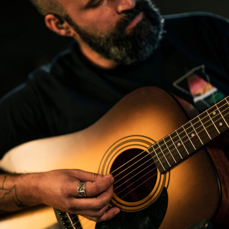 Acoustic guitar being played by a man with a beard, close-up shot with warm lighting.