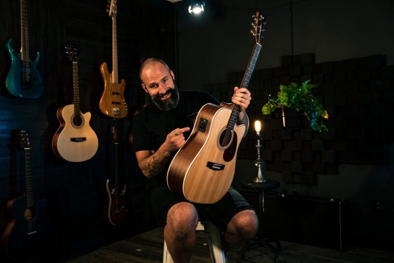 A man with a beard sitting on a stool, holding a wooden acoustic guitar inside a music store with various guitars on the wall behind him.