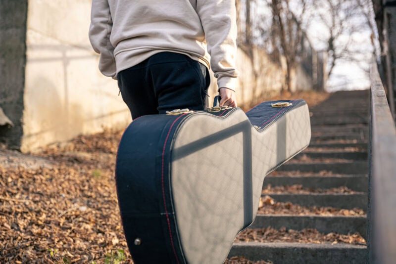 Carrying a black gig bag with red stitching outdoors on stairs, close-up of a musician’s hand holding the case, warm autumn lighting.