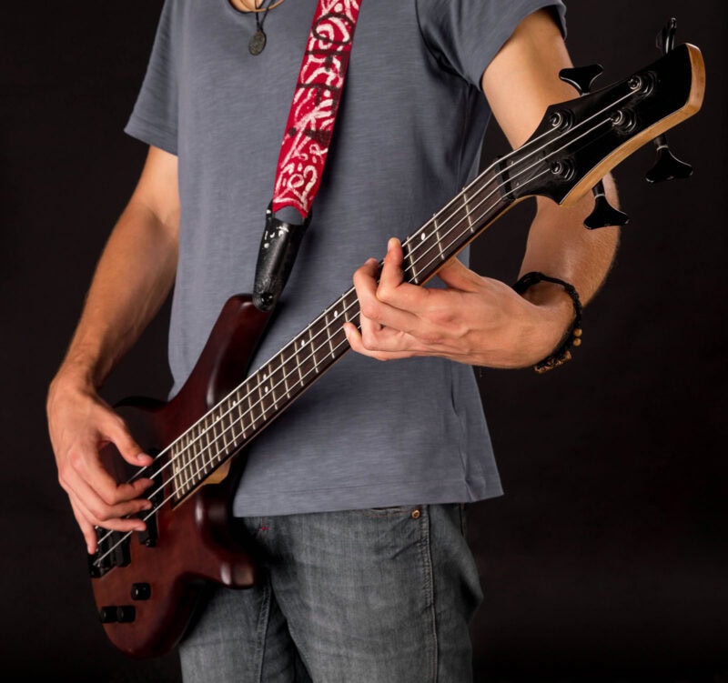 Electric bass guitar being played by a musician wearing a grey t-shirt, with a red patterned strap, against a black background.