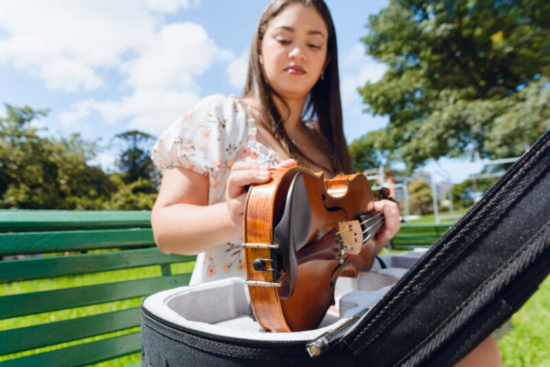 A young woman playing a violin outdoors on a sunny day, sitting on a green park bench.