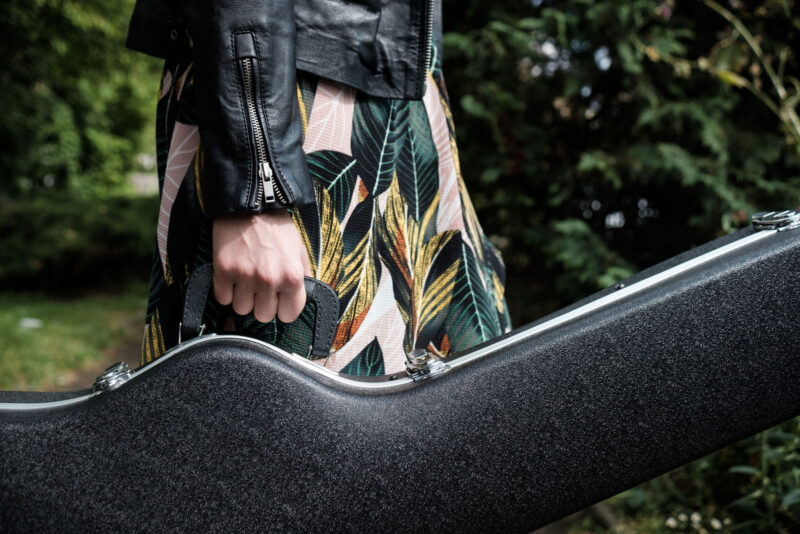 Carrying a black textured guitar case outdoors with a person wearing a colourful floral skirt and a leather jacket.