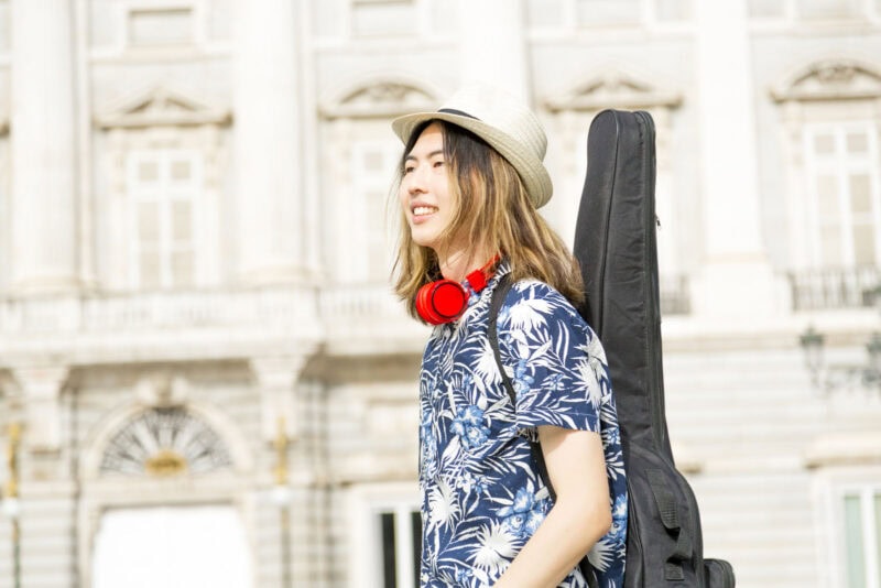 Guitarist with a guitar case on his back, wearing a summer shirt, hat, and headphones, outdoors in front of historic architecture in Dublin.