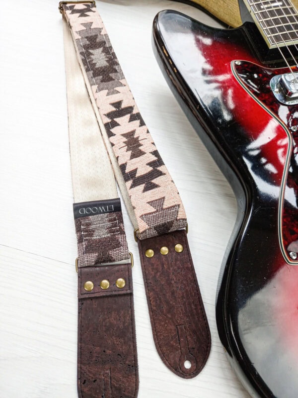 Soft beige and patterned guitar straps with leather ends, laid alongside a red and black electric guitar on a white surface.