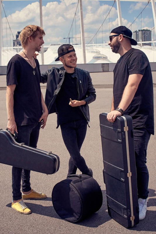 Guitarists carrying instrument cases and chatting outdoors in an urban setting on a sunny day.