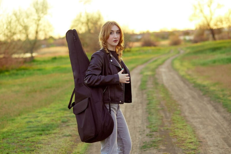 Guitarist carrying a black gig bag over her shoulder outdoors in a scenic rural setting during sunset.
