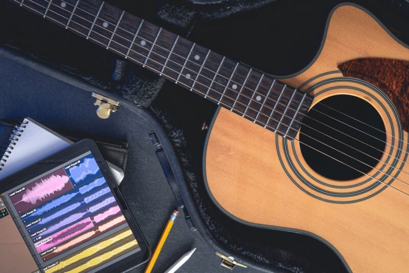 Guitar resting inside its case with an iPad displaying vocal recording tracks and writing tools nearby.